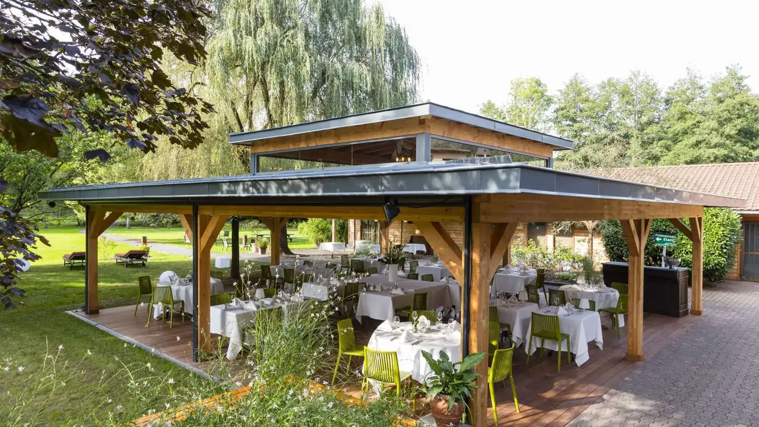 Une terrasse couverte en bois avec des tables dressées pour un repas, entourée d'arbres et de verdure