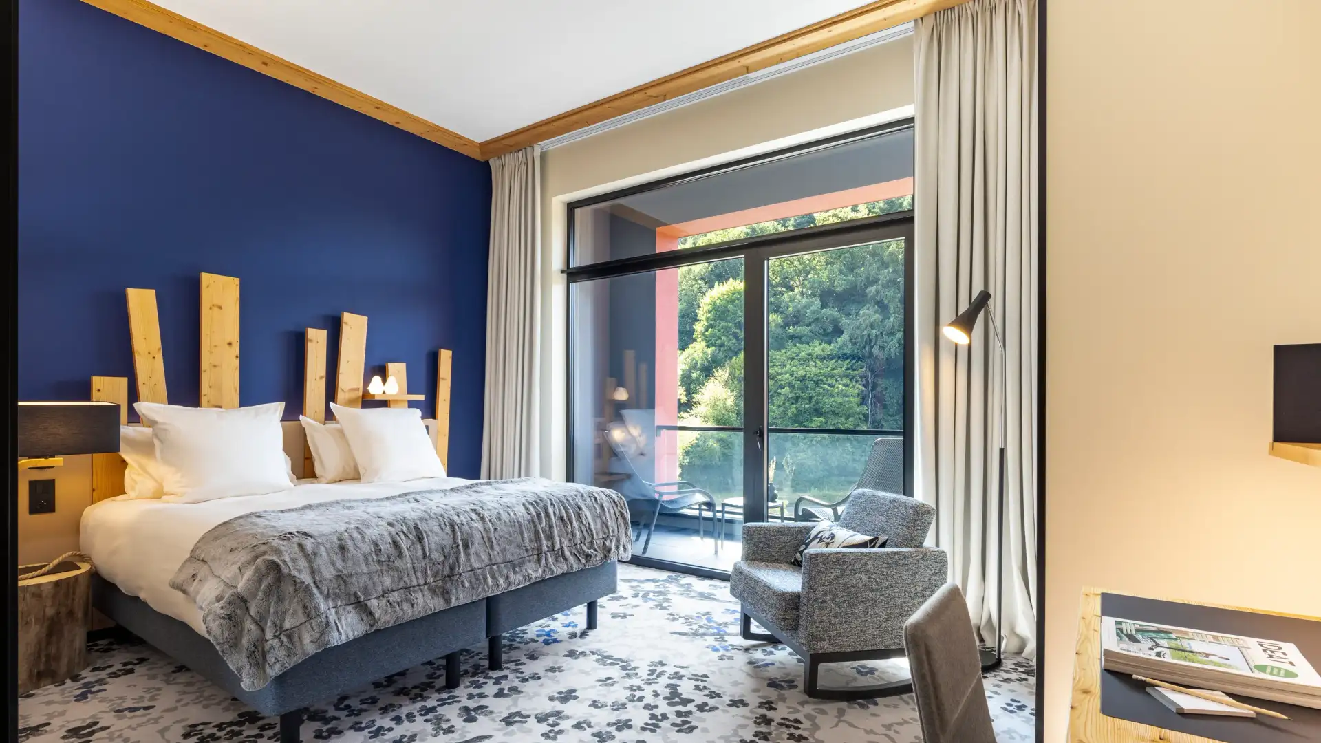Chambre avec un lit double, une table de chevet, un fauteuil et une grande baie vitrée donnant sur une terrasse avec vue sur des arbres