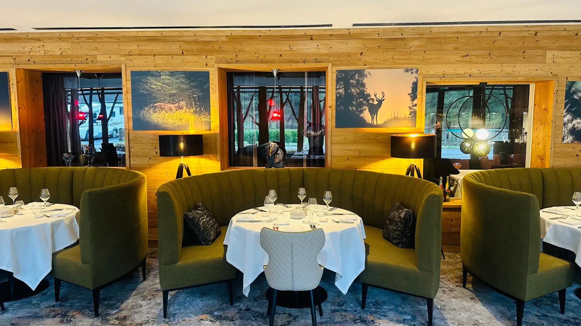 Un restaurant avec des tables dressées, des banquettes vertes et des murs en bois, éclairé par des lampes et des fenêtres avec des rideaux