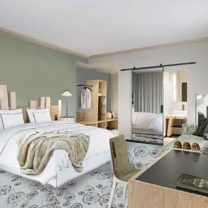 Chambre moderne avec un grand lit blanc, une couverture beige, des tables de chevet en bois, un coin bureau et un dressing visible à travers une porte ouverte