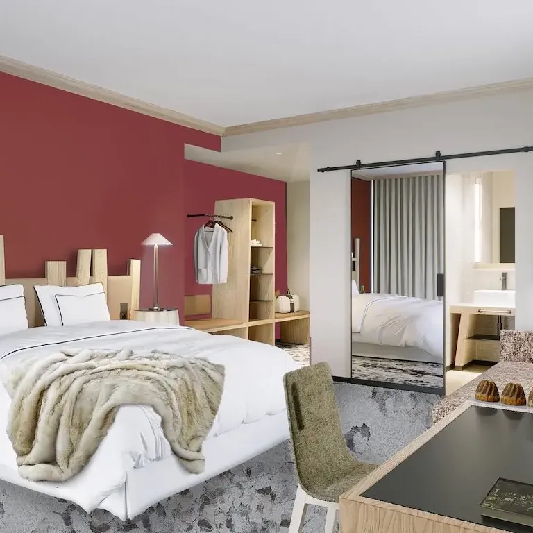 Chambre moderne avec un lit blanc et une couverture en fourrure, des meubles en bois clair, un bureau avec des pâtisseries et une porte ouverte sur une salle de bain
