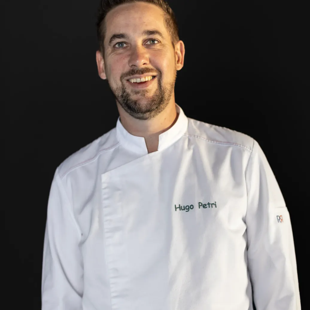 Un homme en veste de chef portant le nom "Hugo Petri" sur la poitrine sourit face à la caméra devant un fond noir