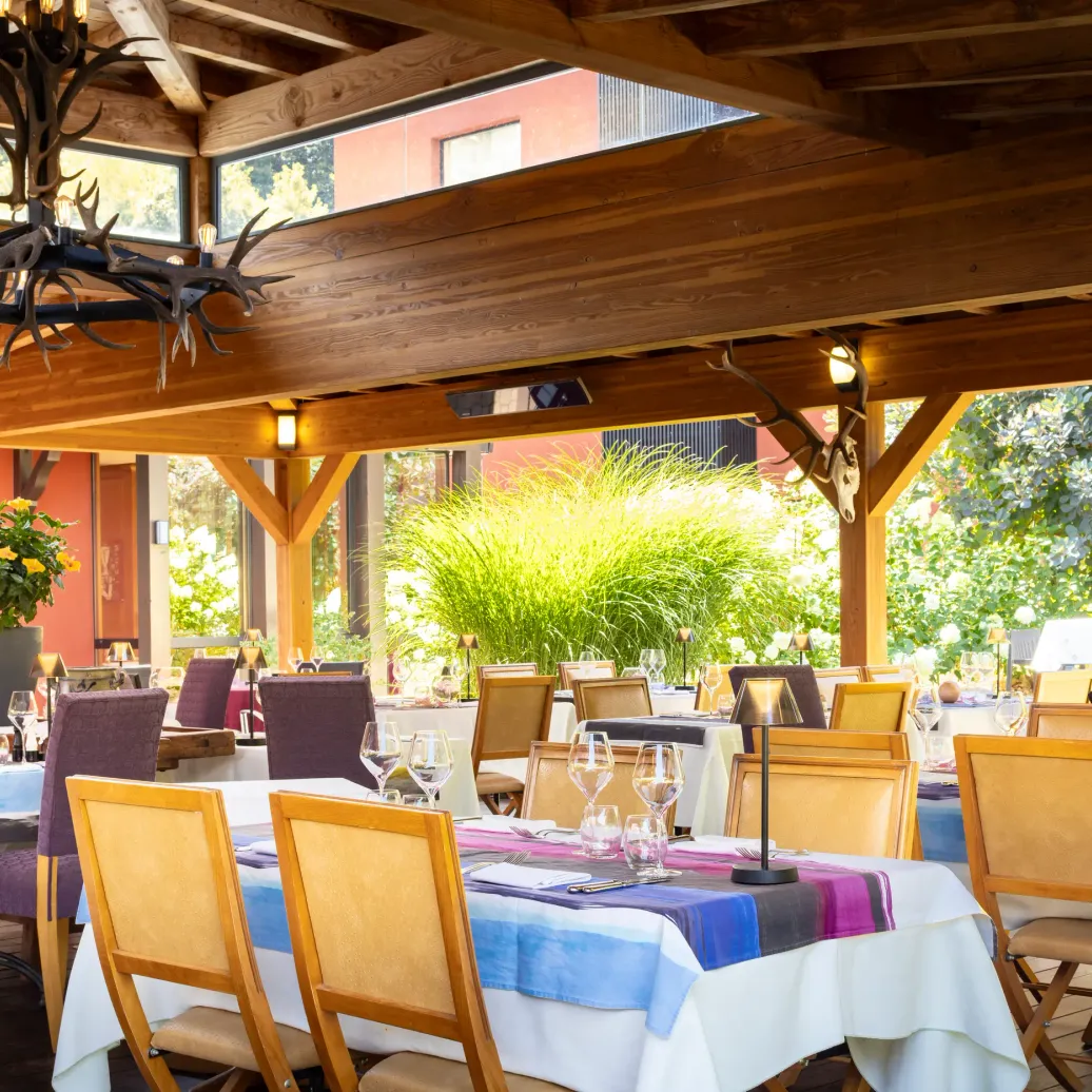 Salle à manger extérieure avec tables dressées, chaises en bois et décorations florales, sous une pergola en bois avec des bois de cerf suspendus