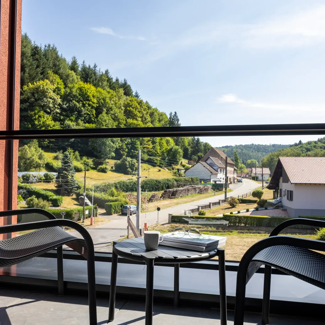 Vue depuis un balcon avec table, chaises et tasse, sur un village avec maisons et verdure en arrière-plan