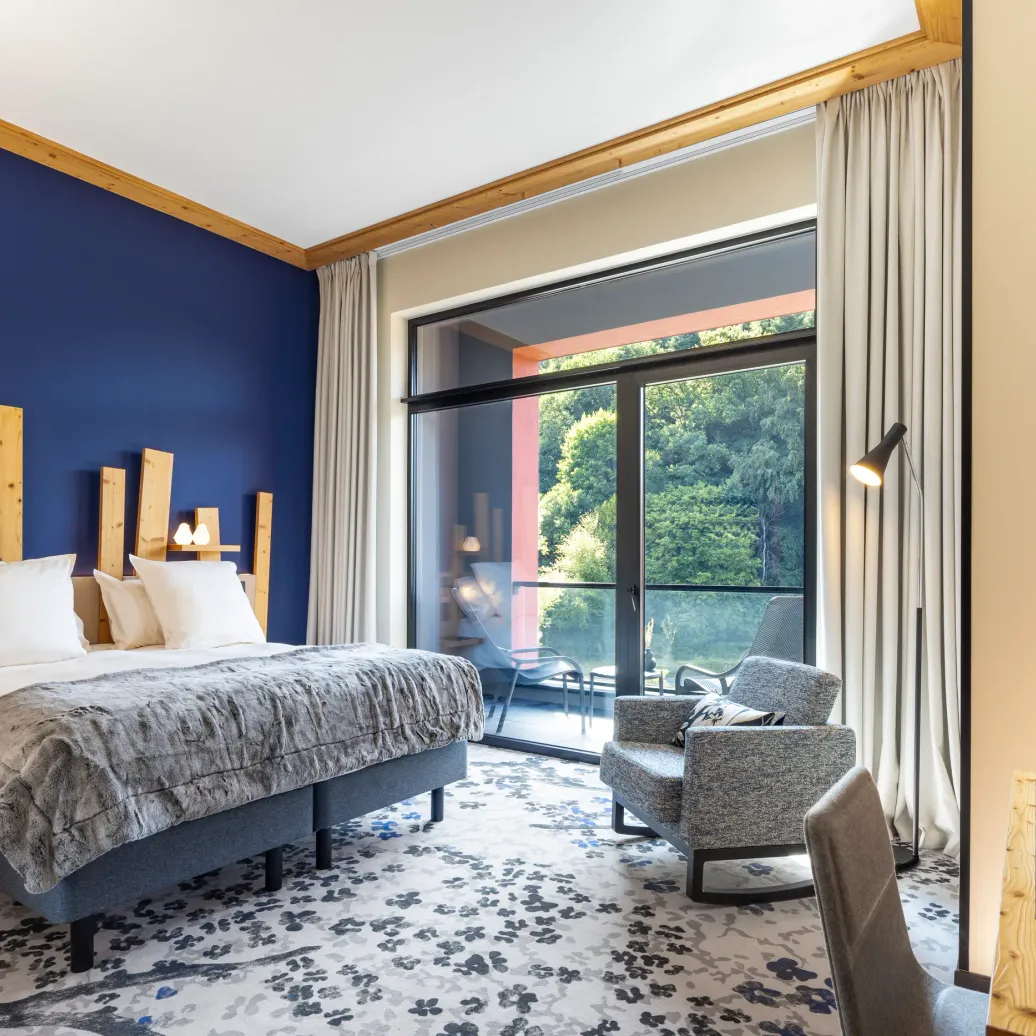 Chambre avec un lit double, une table de chevet, un fauteuil et une grande baie vitrée donnant sur une terrasse avec vue sur des arbres