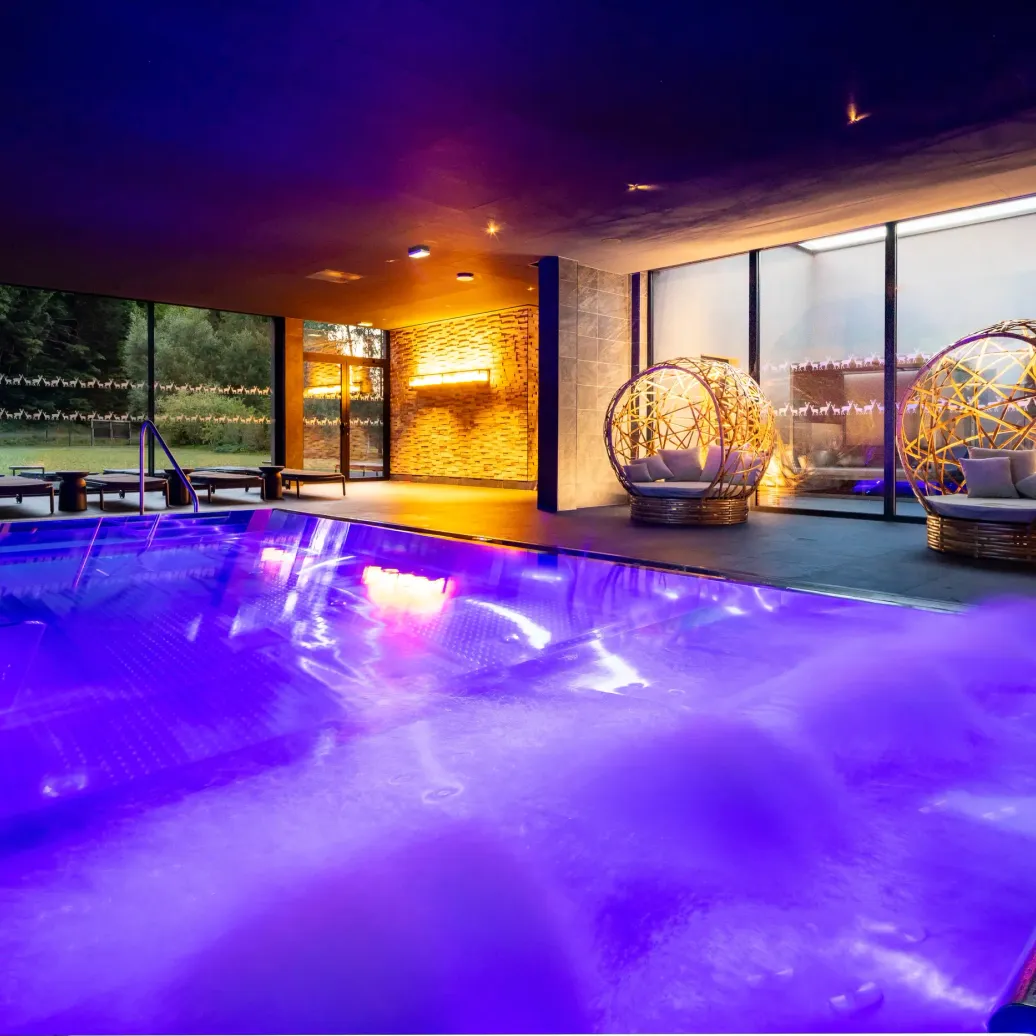 Piscine intérieure éclairée en violet avec des chaises longues et des fauteuils sphériques en osier illuminés à proximité, entourée de grandes baies vitrées donnant sur un jardin
