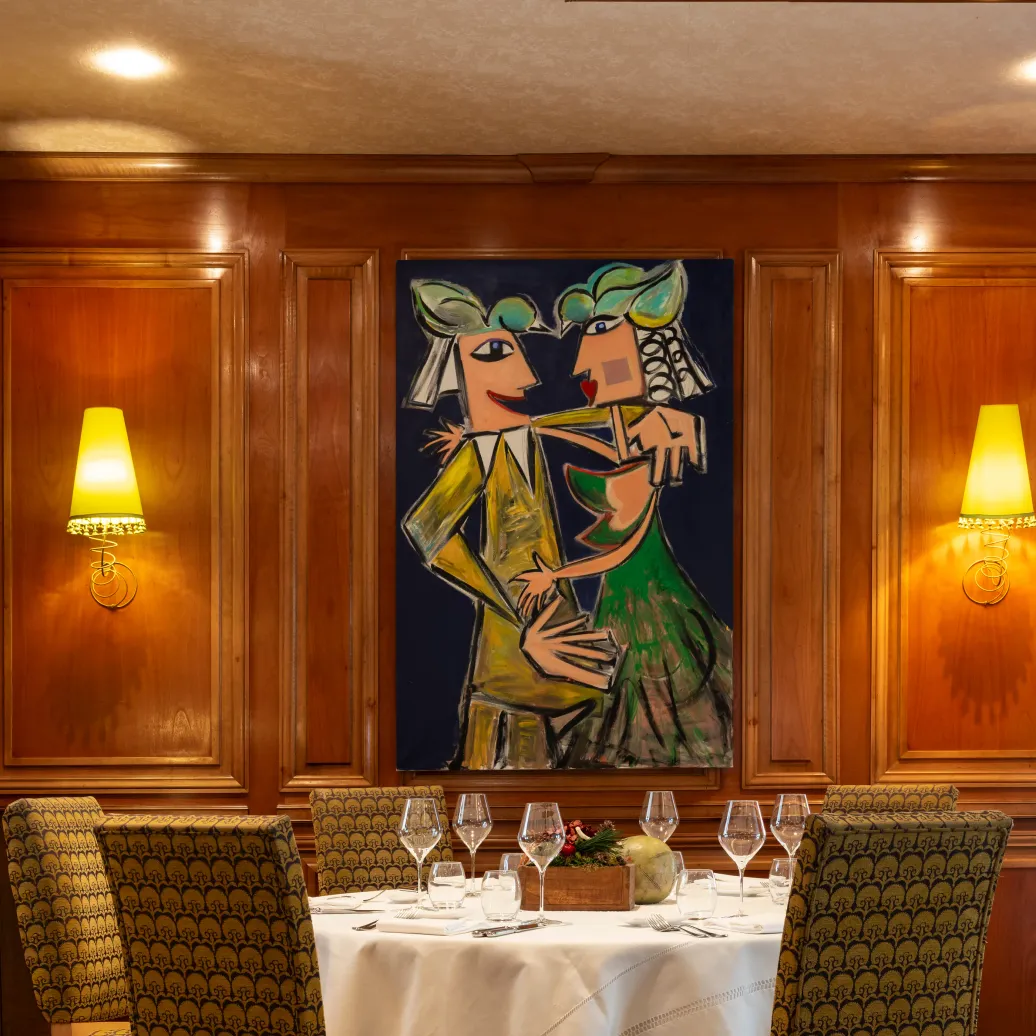Table dressée avec verres et couverts, entourée de chaises à motifs, dans une salle à manger avec un tableau mural représentant un couple et des vitraux colorés au plafond