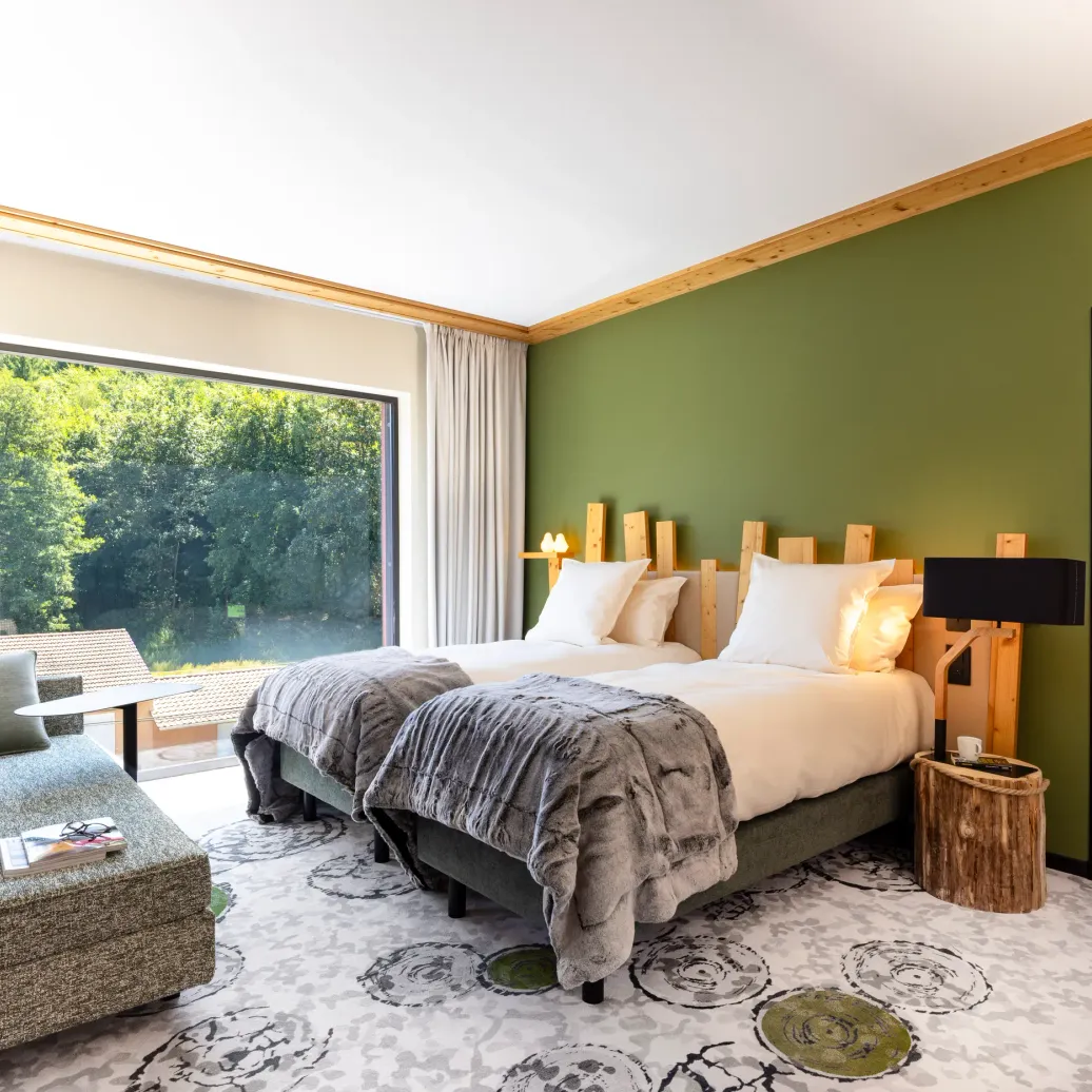 Chambre avec deux lits, une chaise longue, une table de chevet en bois et une grande fenêtre donnant sur une forêt