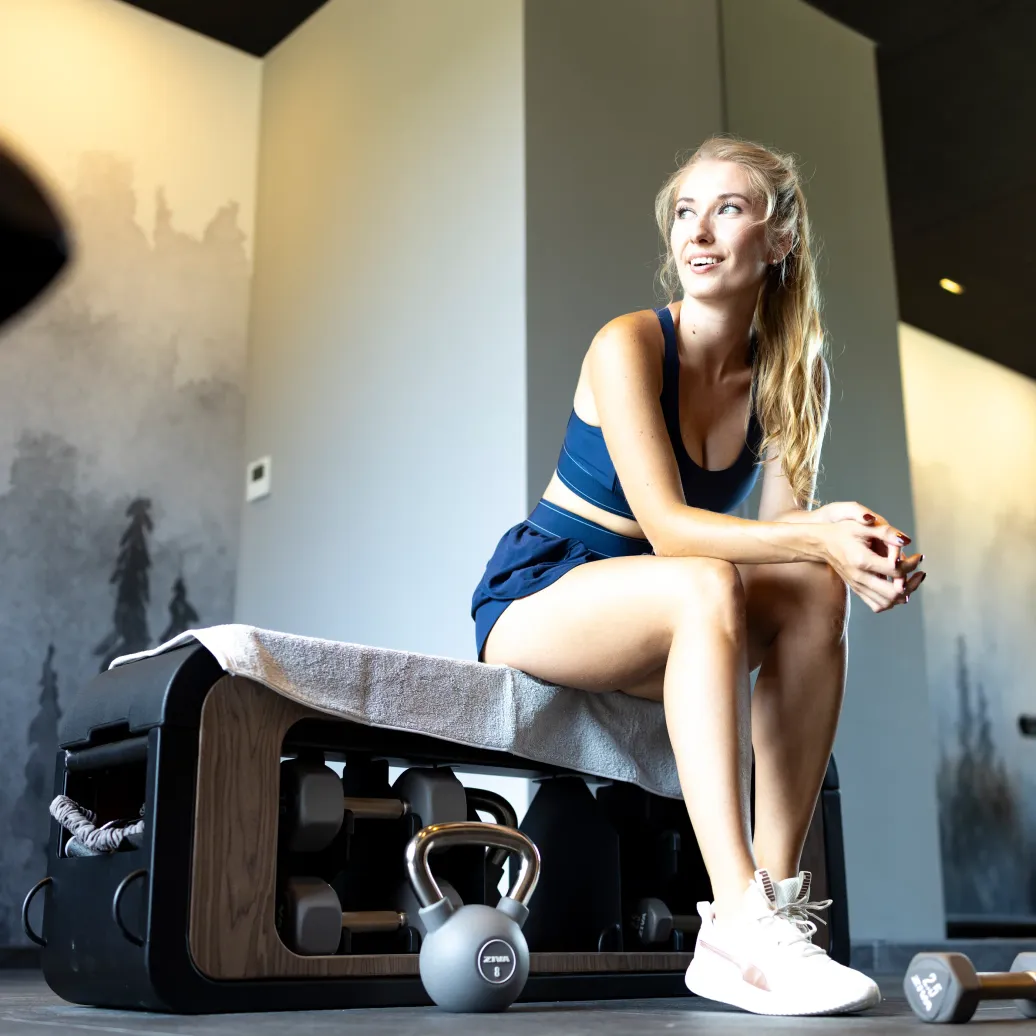 Une femme assise sur un banc de musculation avec une serviette, entourée de matériel de fitness dans une salle de sport