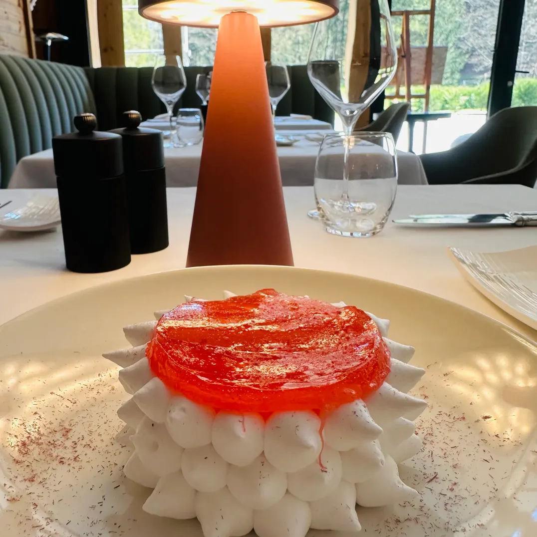 Un dessert composé de meringues empilées recouvertes d'une gelée rouge, posé sur une assiette blanche avec des éclats de chocolat, dans un restaurant avec des tables et des chaises visibles en arrière-plan