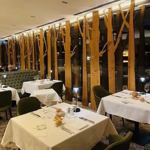 Restaurant avec tables dressées, nappes blanches et chaises vertes, devant une paroi décorative en bois imitant des troncs d'arbres