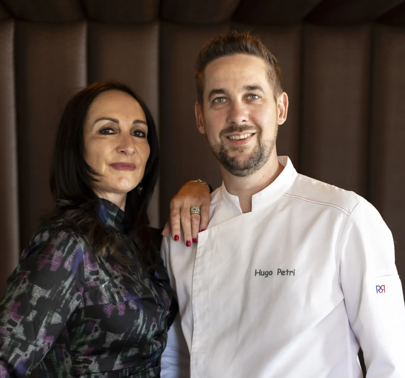 Deux personnes posent côte à côte, l'une en tenue de chef avec le nom "Hugo Petri" brodé, l'autre en robe à motifs avec une écharpe.