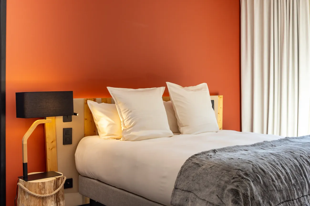 Lit avec deux oreillers blancs, une lampe murale en bois et une couverture grise, devant un mur orange et une fenêtre avec des rideaux blancs