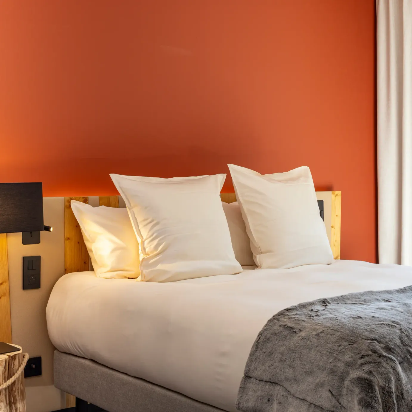 Bett mit zwei weißen Kissen, einer Holz-Wandleuchte und einer grauen Decke, vor einer orangen Wand und einem Fenster mit weißen Vorhängen