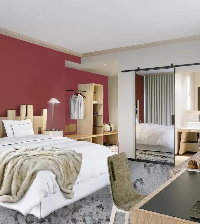 Chambre moderne avec un lit blanc et une couverture en fourrure, des meubles en bois clair, un bureau avec des pâtisseries et une porte ouverte sur une salle de bain