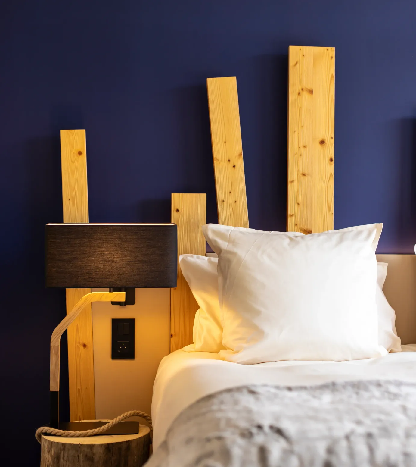 Lit avec une tête de lit en bois clair, deux oreillers blancs et une couverture grise, une lampe de chevet noire et un panier en bois à côté