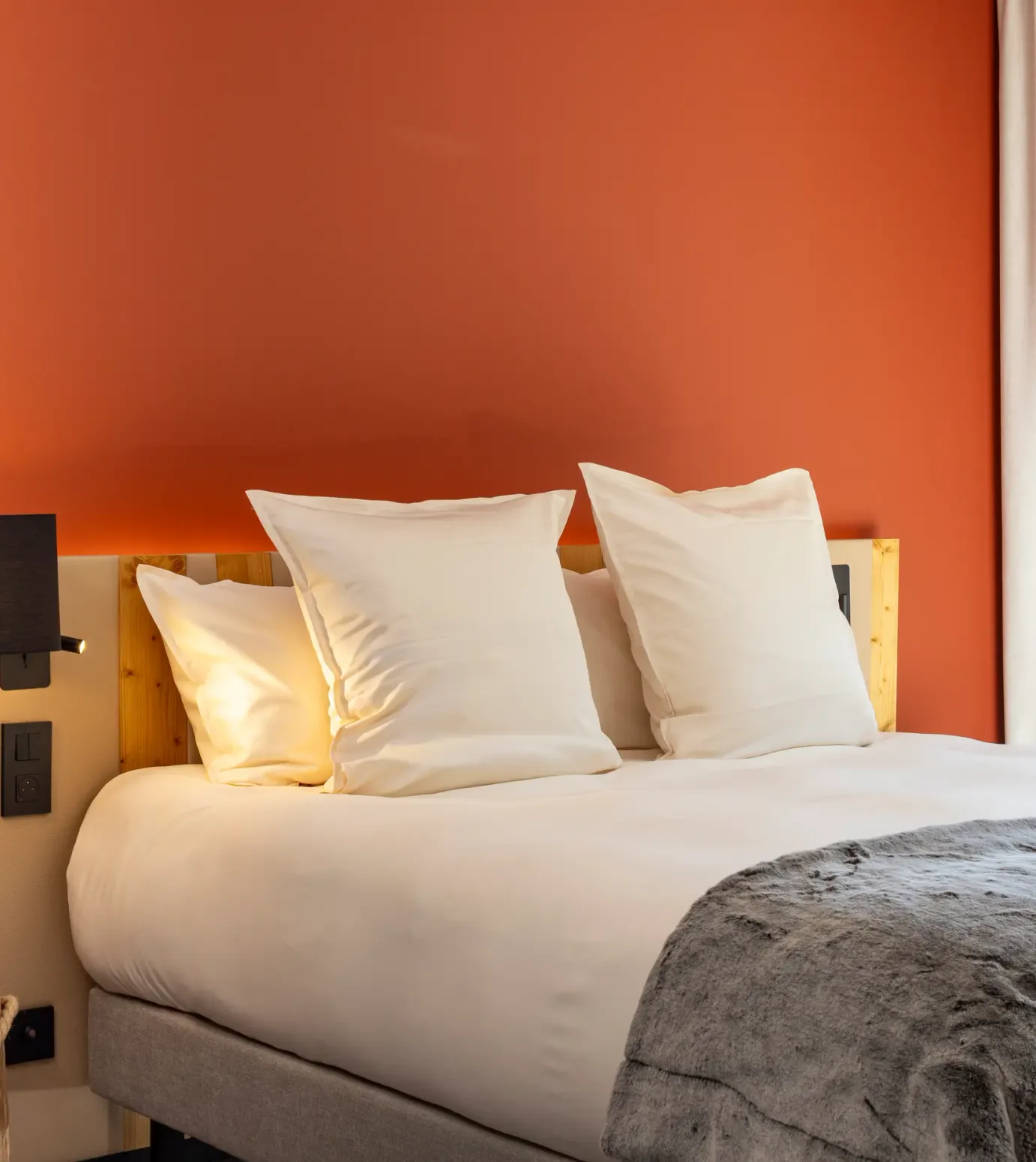 Lit avec deux oreillers blancs, une lampe murale en bois et une couverture grise, devant un mur orange et une fenêtre avec des rideaux blancs