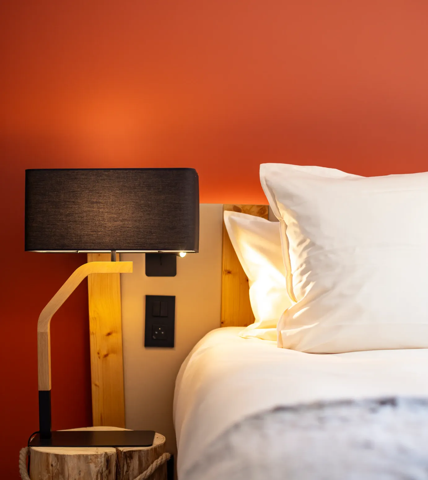 Lit avec une tête de lit en bois clair, deux oreillers blancs et une lampe de chevet à abat-jour noir sur une table de nuit en bois brut