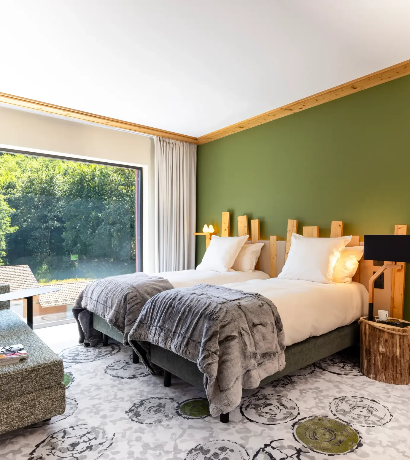 Chambre avec deux lits, une chaise longue, une table de chevet en bois et une grande fenêtre donnant sur une forêt