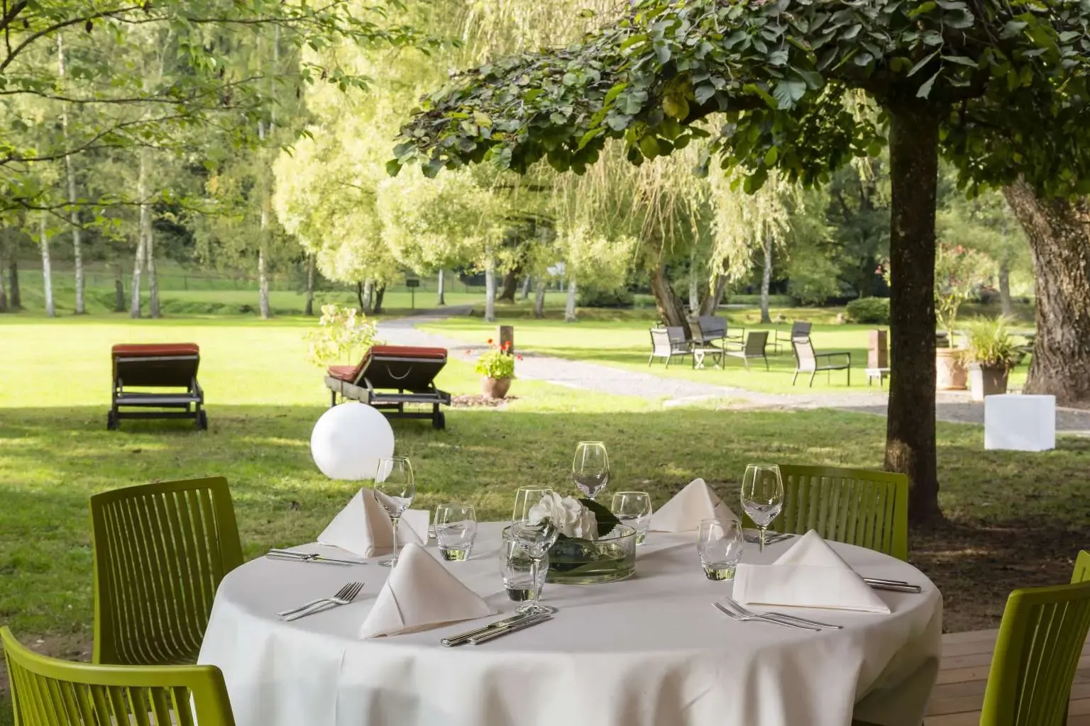 Table dressée en extérieur sous des arbres avec des verres, des couverts et des serviettes, entourée de chaises vertes