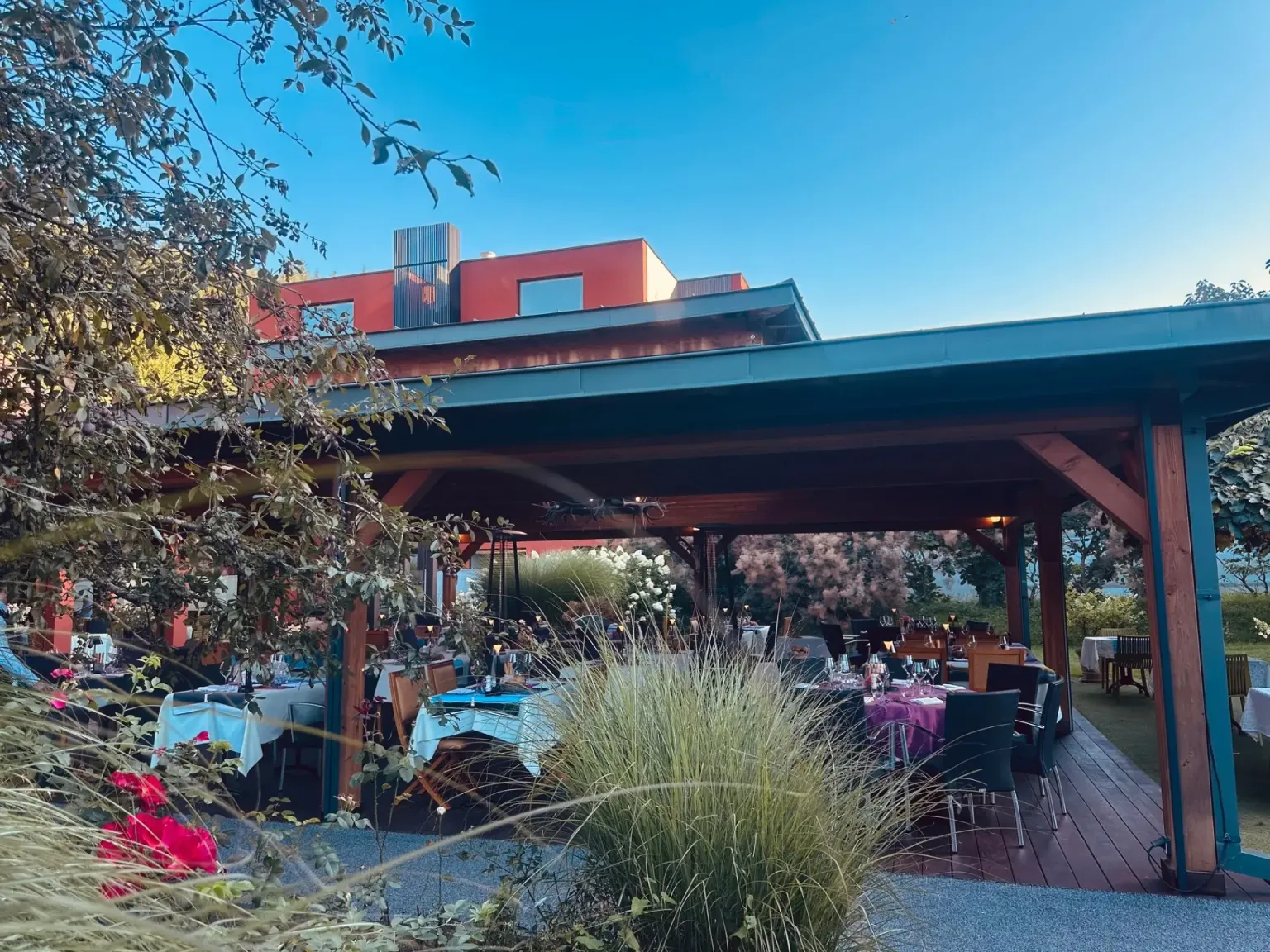 Une terrasse couverte avec des tables dressées pour un repas, entourée de végétation et de fleurs, sous un ciel dégagé