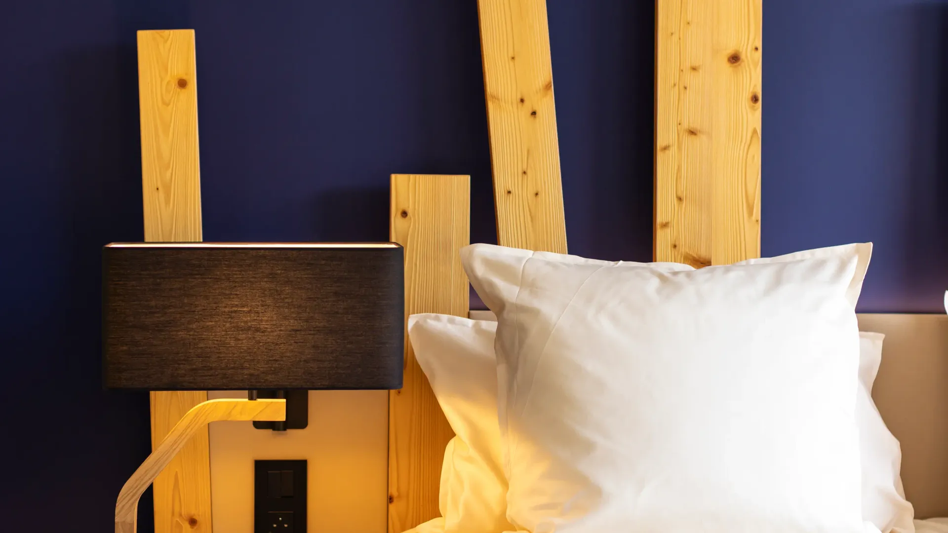 Lit avec une tête de lit en bois clair, deux oreillers blancs et une couverture grise, une lampe de chevet noire et un panier en bois à côté
