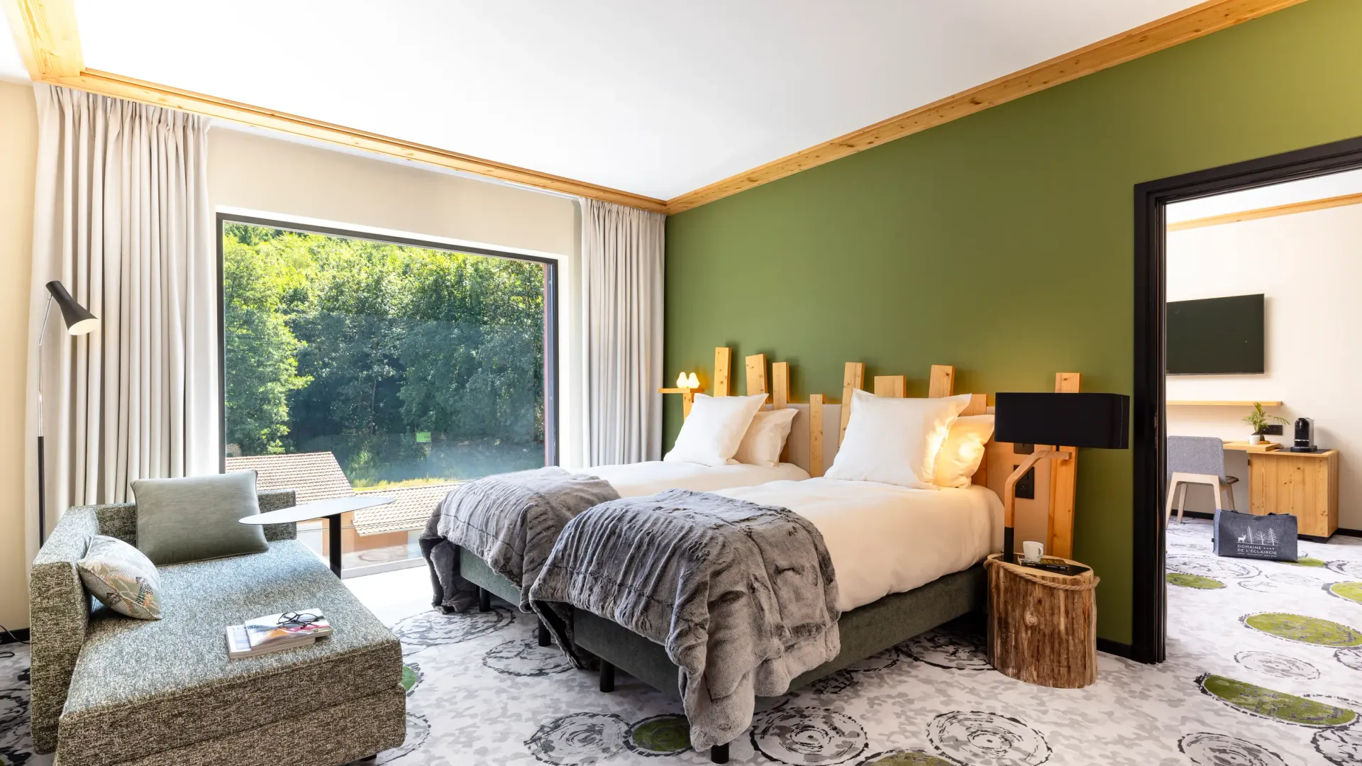 Chambre avec deux lits, une chaise longue, une table de chevet en bois et une grande fenêtre donnant sur une forêt