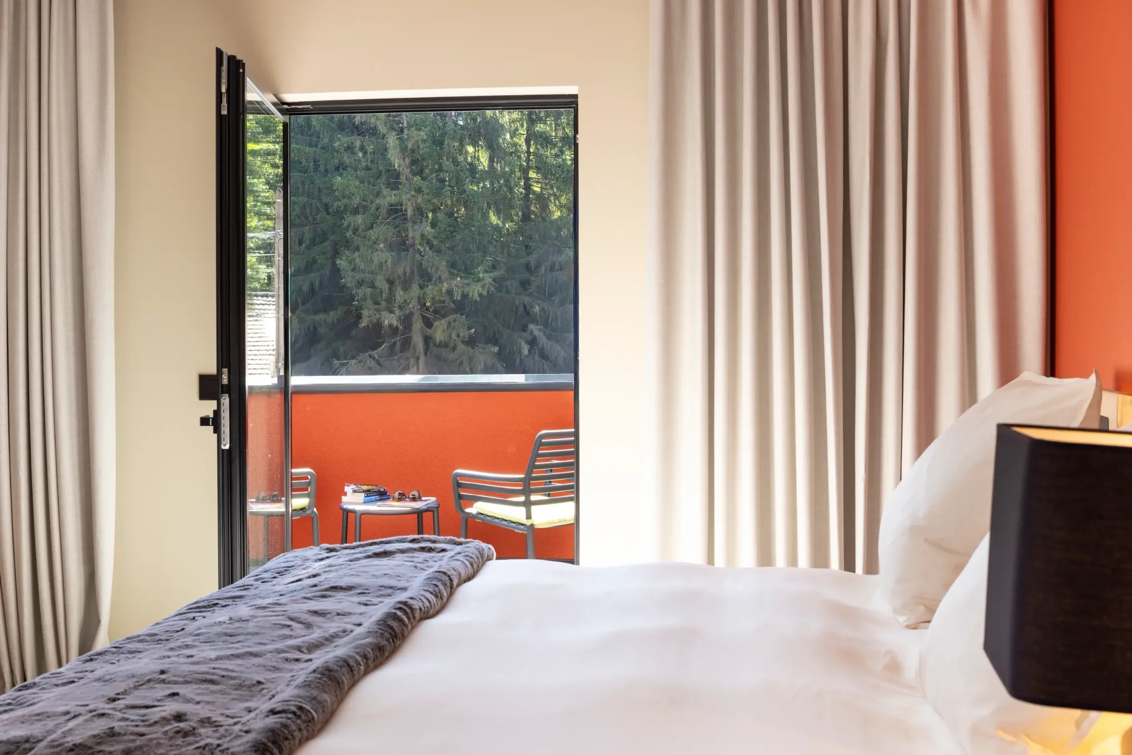 Zimmer mit einem Bett und einer offenen Glastür zum Balkon mit Stühlen und einem Tisch, Blick auf Bäume
