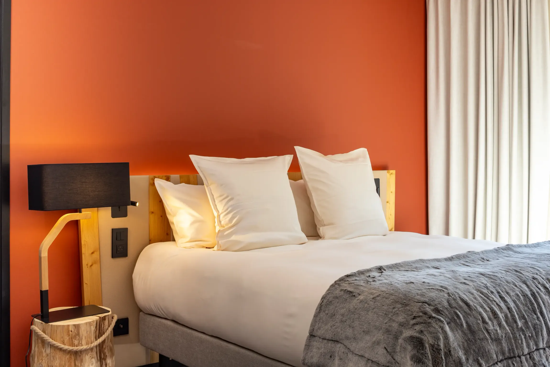 Bed with two white pillows, a wooden wall lamp, and a gray blanket, in front of an orange wall and a window with white curtains.