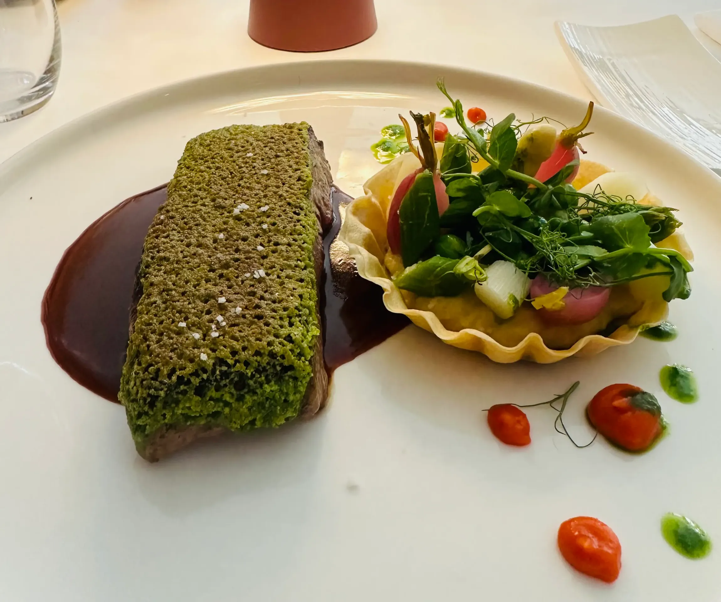 Un plat composé d'un rectangle vert croustillant sur une sauce brune accompagné d'une salade de jeunes pousses et de tomates cerises
