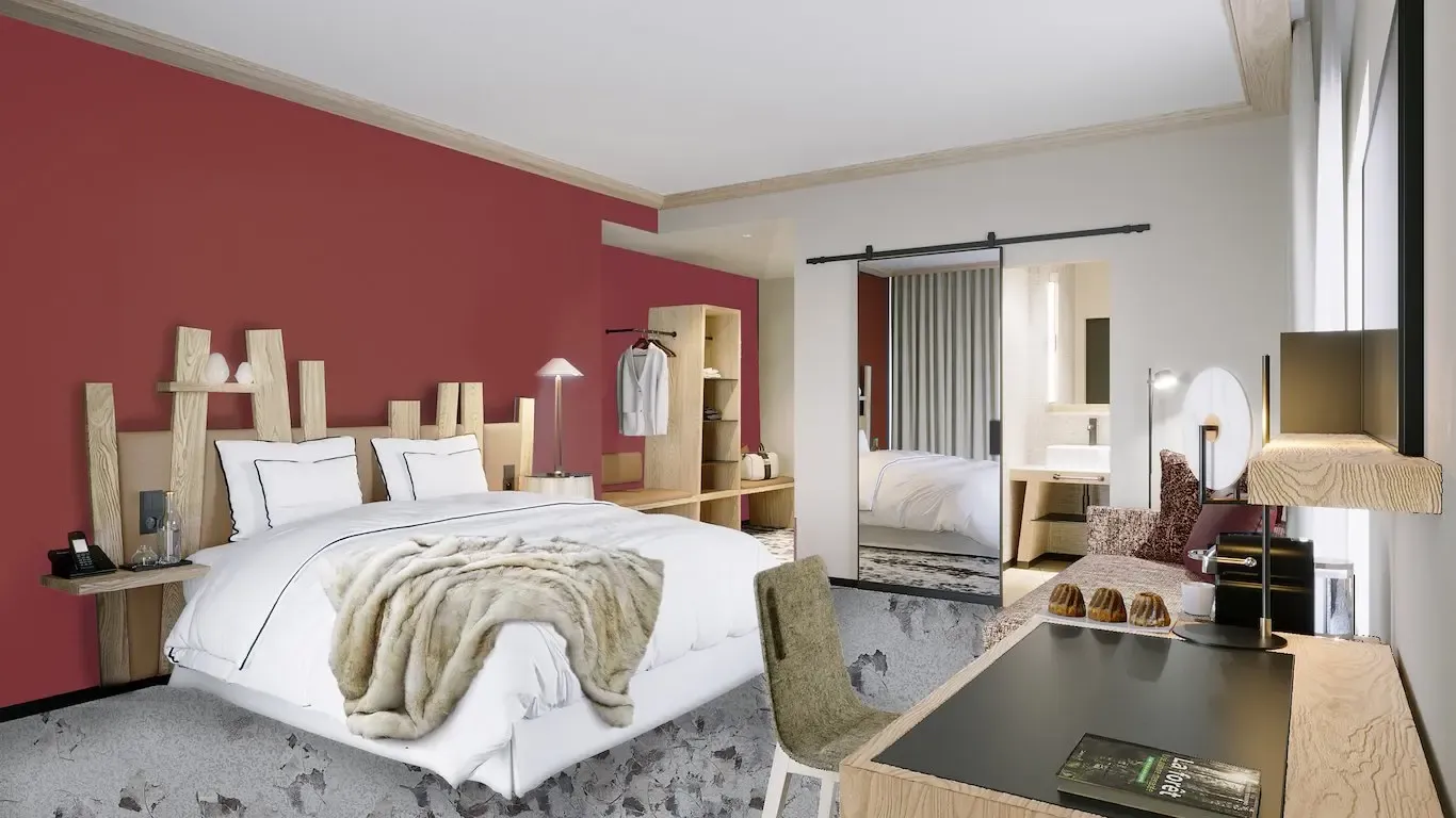 Chambre moderne avec un lit blanc et une couverture en fourrure, des meubles en bois clair, un bureau avec des pâtisseries et une porte ouverte sur une salle de bain