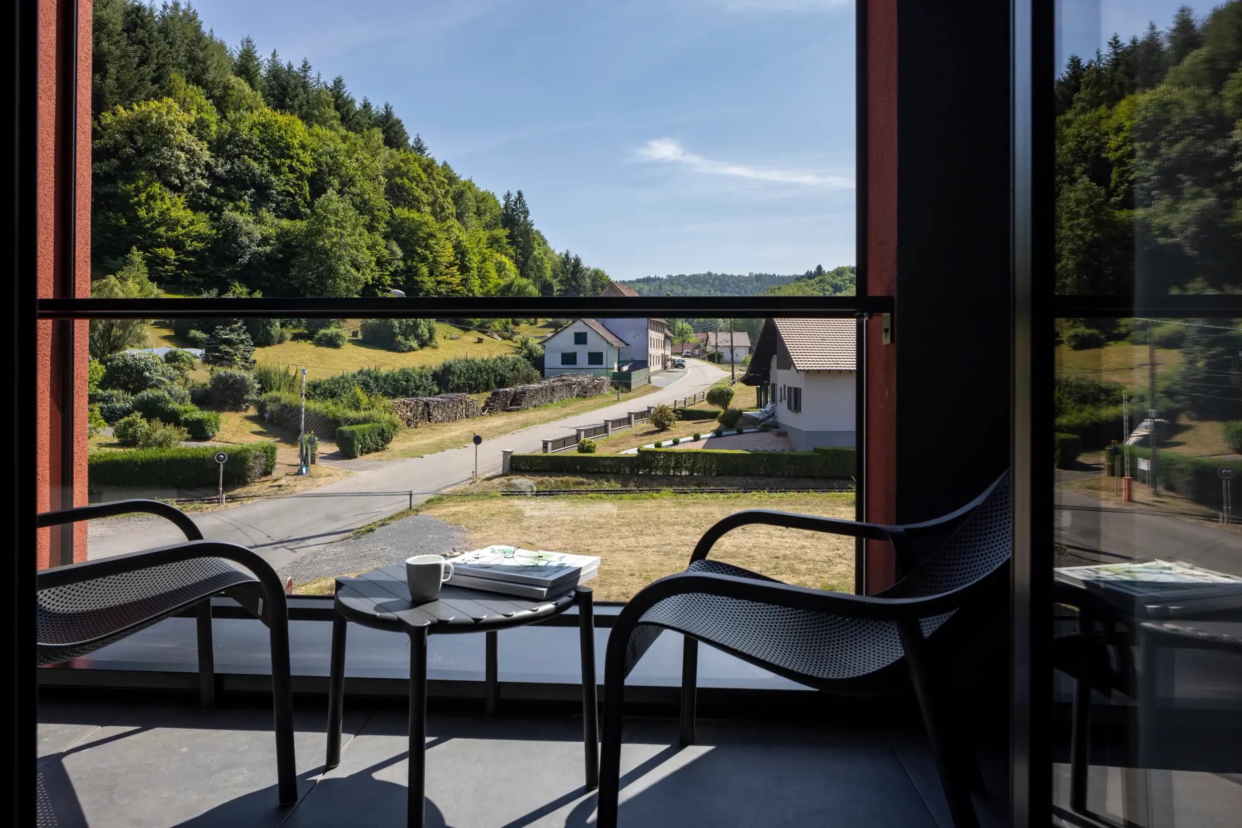 Vue depuis une fenêtre ouverte sur un balcon avec des chaises et une table, montrant une route de campagne bordée de maisons et de verdure sous un ciel dégagé
