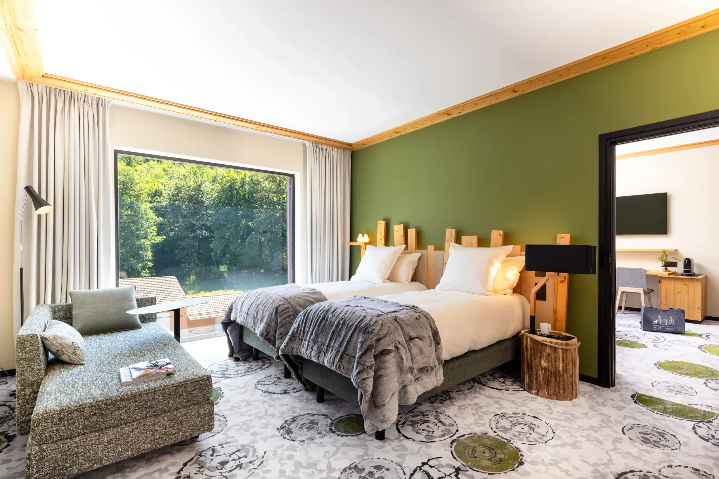 Room with two beds, a sun lounger, a wooden bedside table and a large window overlooking a forest