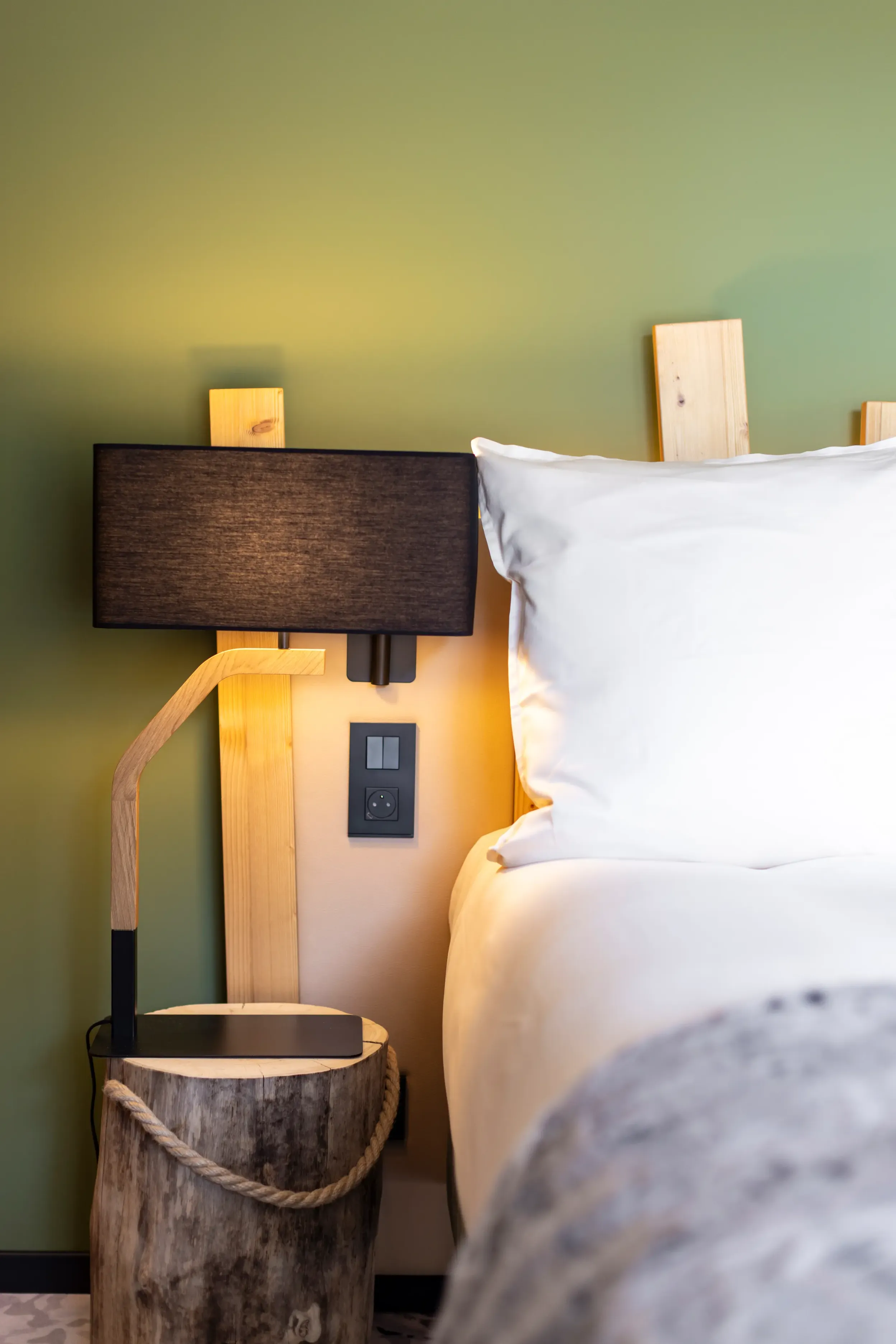 Une lampe sur pied en bois avec un abat-jour noir est placée à côté d'un lit avec une tête de lit en bois clair et des oreillers blancs