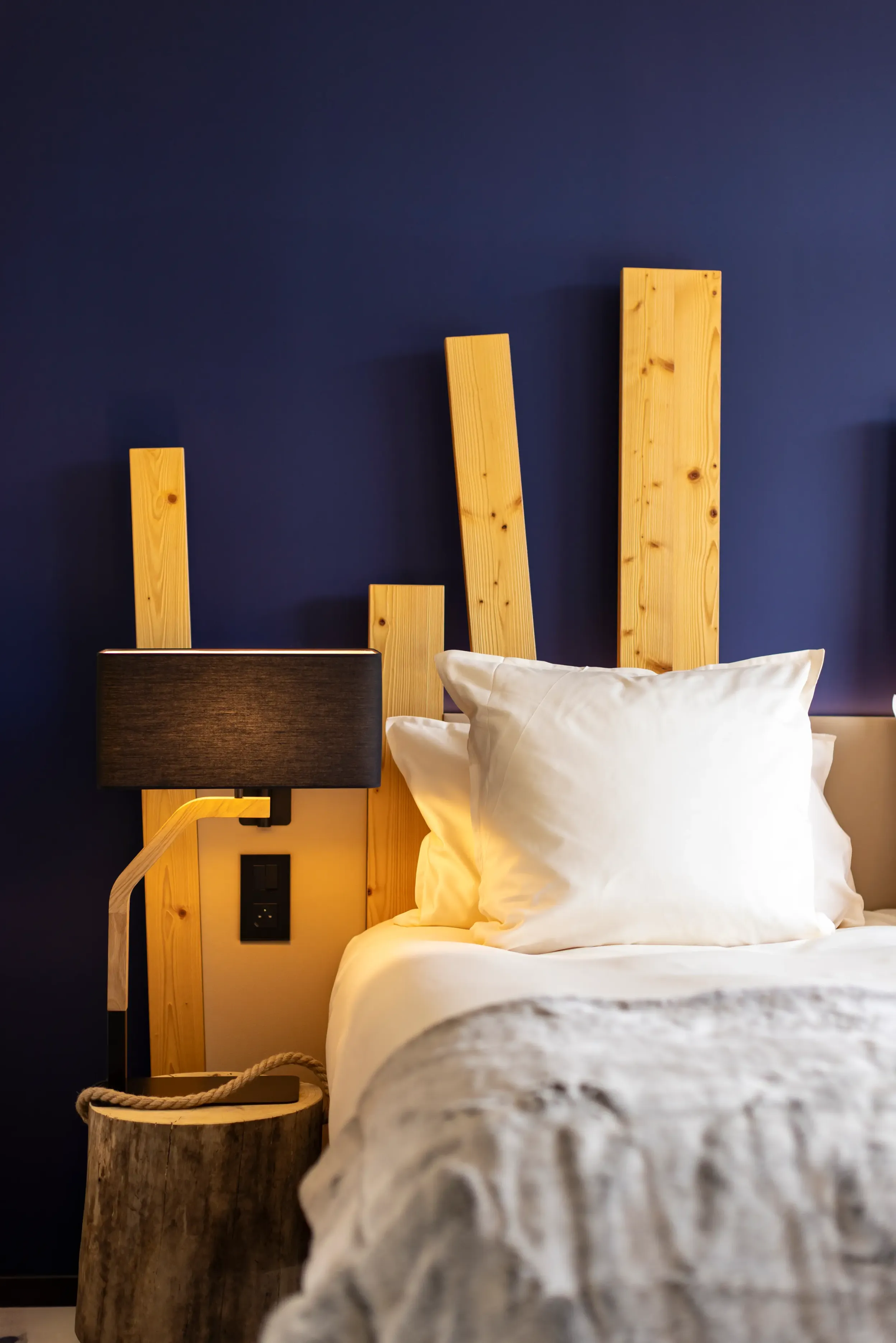 Lit avec une tête de lit en bois clair, deux oreillers blancs et une couverture grise, une lampe de chevet noire et un panier en bois à côté