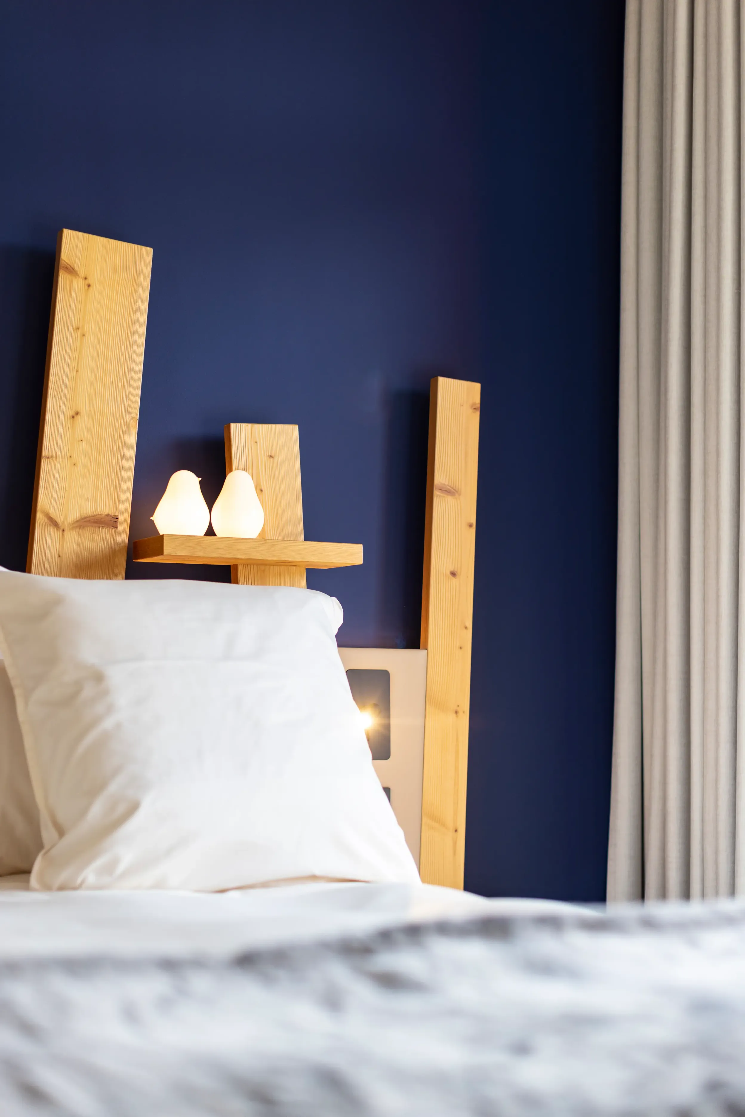 Un lit avec une tête de lit en bois clair et deux bougies allumées posées sur une étagère en bois, contre un mur bleu foncé