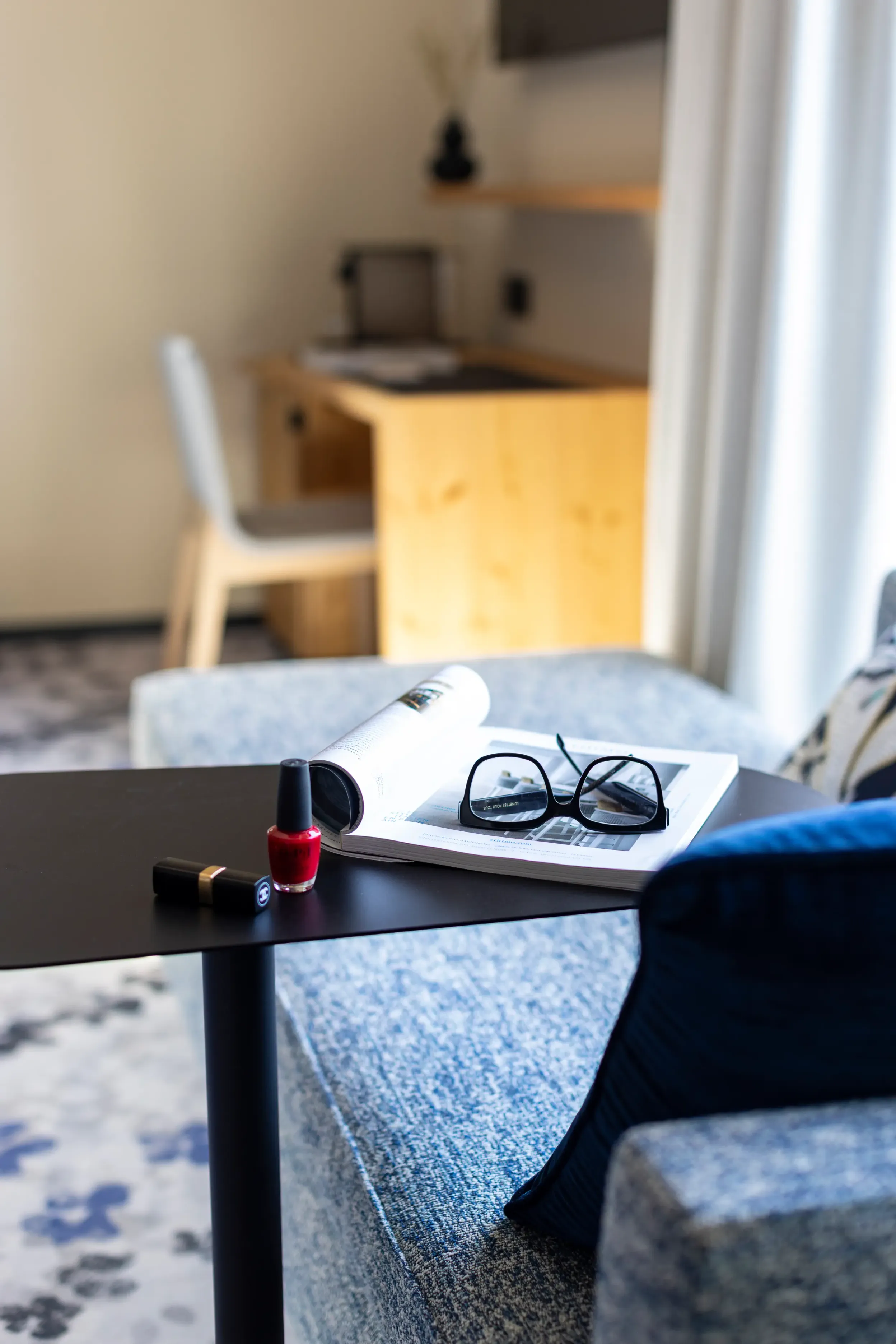 Une table basse avec une bouteille de vernis à ongles rouge, des lunettes et un magazine ouvert, dans un salon avec un canapé et une table en bois en arrière-plan