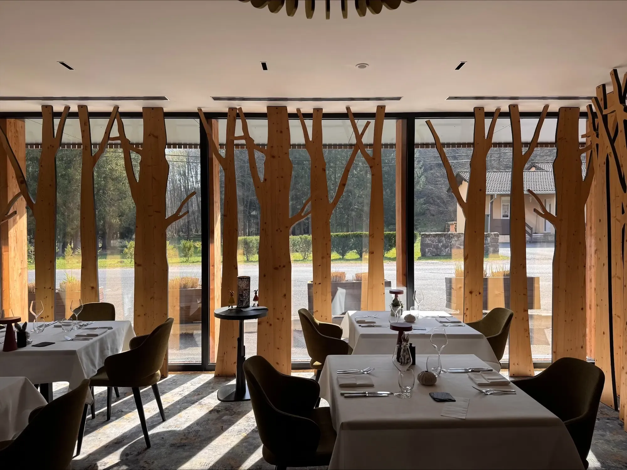 Restaurant avec tables dressées, grandes baies vitrées et colonnes en bois imitant des arbres, vue sur l'extérieur