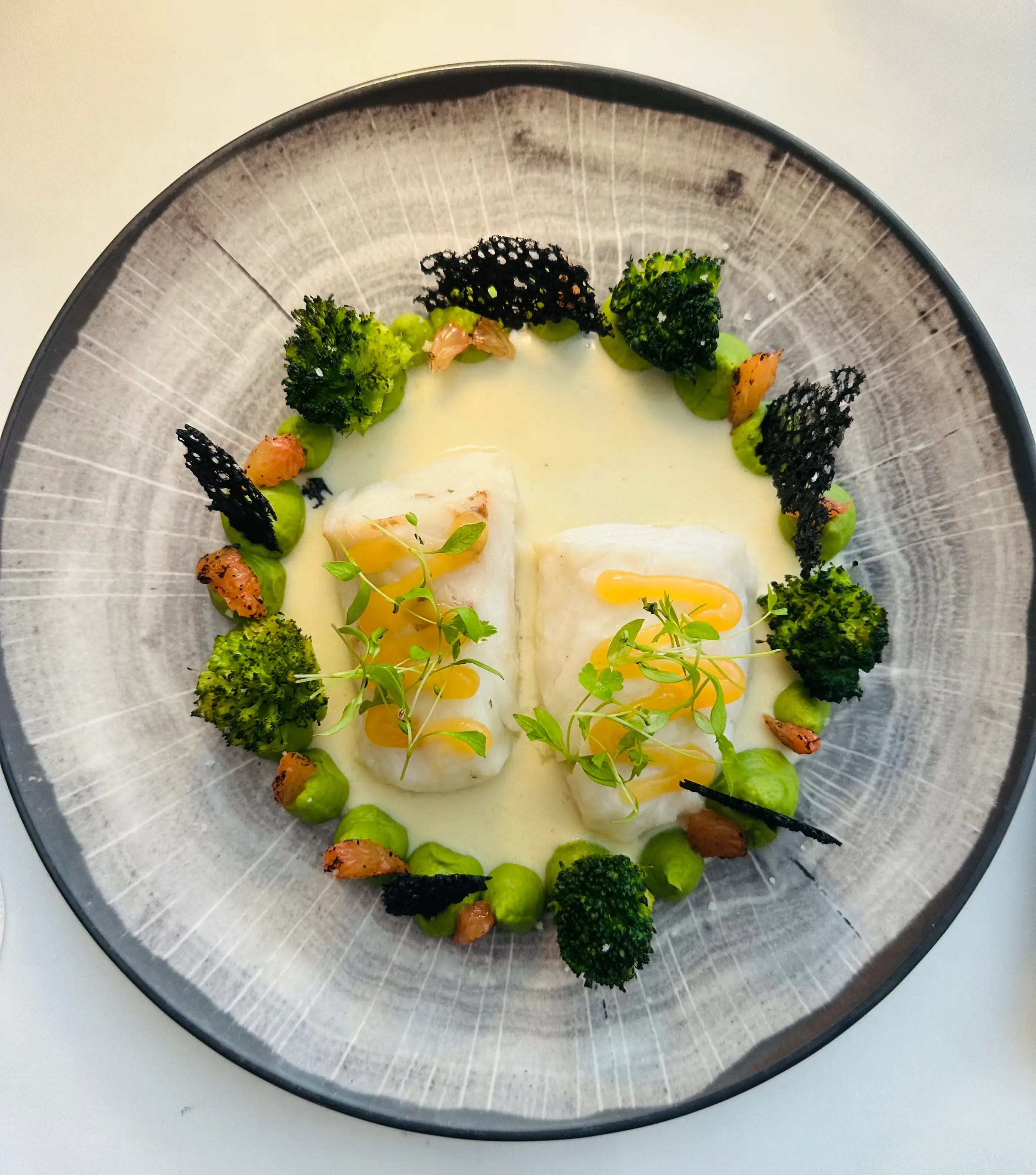 Un plat composé de deux morceaux de poisson blanc, de brocolis, de sauce blanche et de garnitures diverses sur une assiette grise