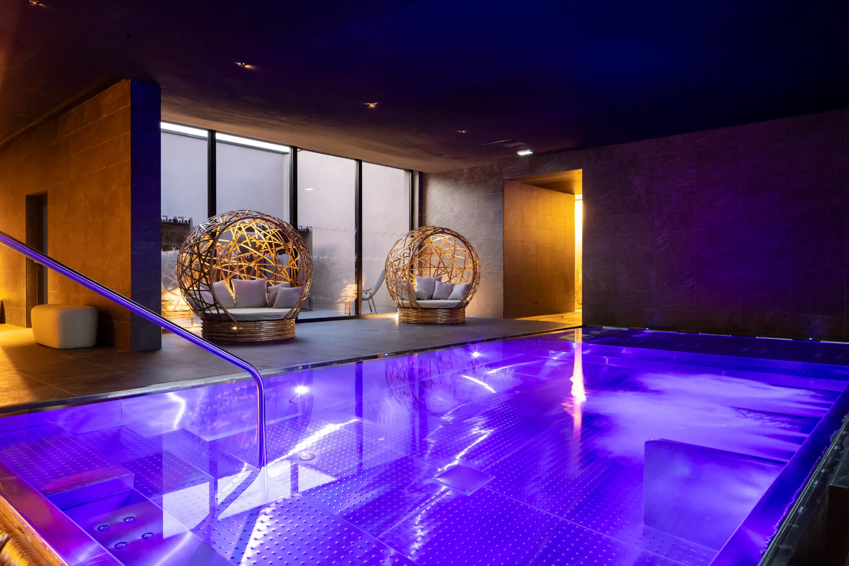 Piscine intérieure éclairée en violet avec des sphères en osier servant de sièges dans un espace moderne aux murs sombres