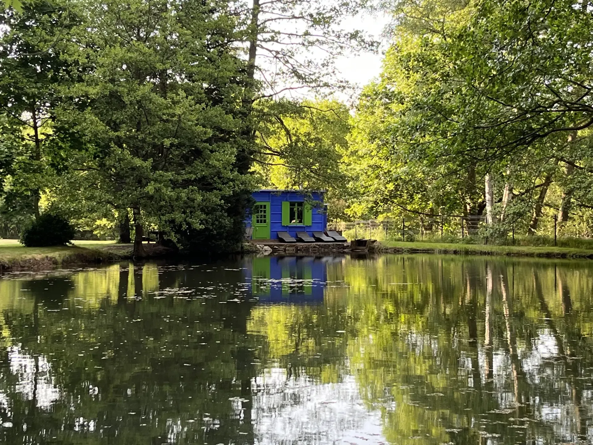 Une petite maison bleue avec des volets verts est située au bord d'un étang entouré d'arbres