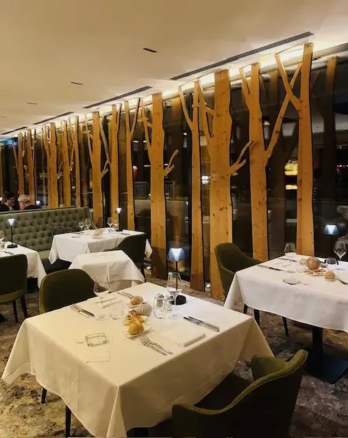 Restaurant avec tables dressées, nappes blanches et chaises vertes, devant une paroi décorative en bois imitant des troncs d'arbres